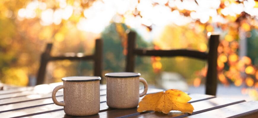 Two mugs sitting on an outdoor wooden table with fall foliage in the background